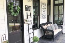 a modern farmhouse porch with a black wicker sofa with lots of pillows, a ladder, some signs and a greenery wreath