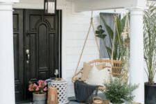a modern front porch with a rattan suspended bench, potted greenery and blooms, candle lanterns and a black pendant lamp