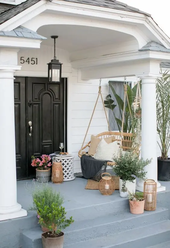 a modern front porch with a rattan suspended bench, potted greenery and blooms, candle lanterns and a black pendant lamp