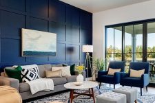 a modern living room with a navy paneled wall, chic grey and navy furniture, a tan sofa, a printed rug and blankets