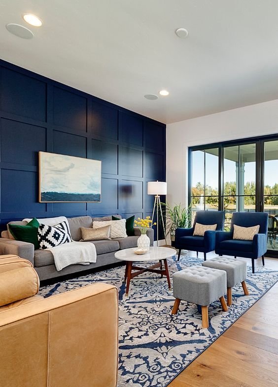 a modern living room with a navy paneled wall, chic grey and navy furniture, a tan sofa, a printed rug and blankets