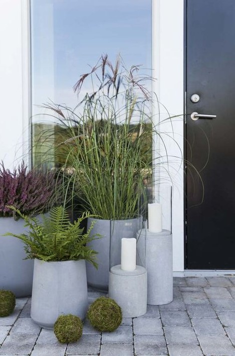 a modern porch with a black door, concrete planters with various plants, candle lanterns and moss balls for decor