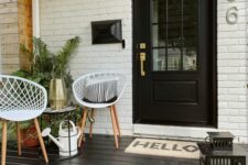 a modern porch with a black floor, white chairs, a side table with a gold sconce, a white watering can and some greenery