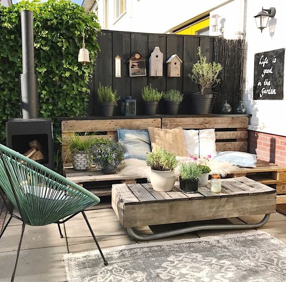 a modern rustic terrace with a pallet bench, pretty textiles, a pallet coffee table, a green chair and a hearth plus potted greenery