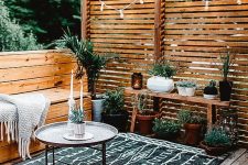 a modern rustic terrace with a wooden deck, built-in benches with a blanket, a bench with potted plants and some lights over the space