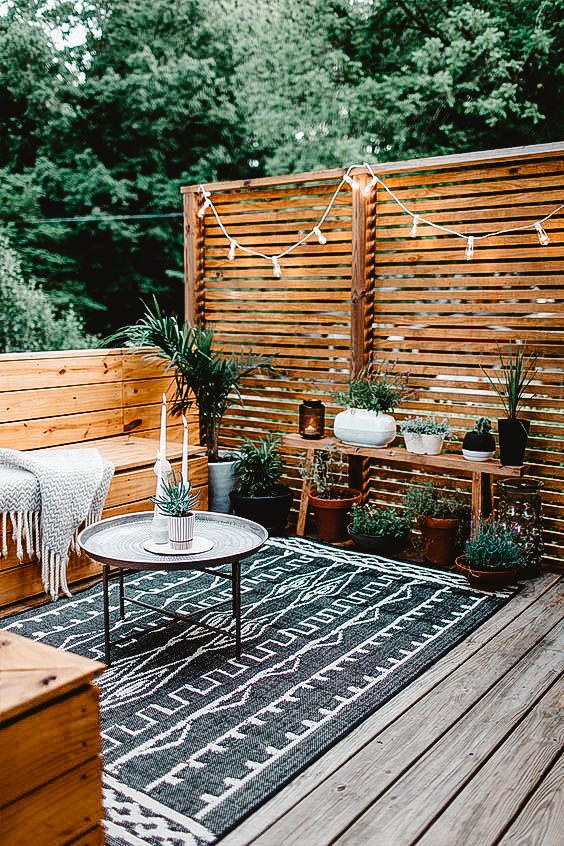 A modern rustic terrace with a wooden deck, built in benches with a blanket, a bench with potted plants and some lights over the space