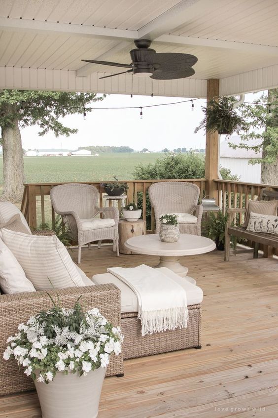 a neutral farmhouse porch with a wooden deck, wicker furniture, potted plants and blooms, a bench and tree stump side tables