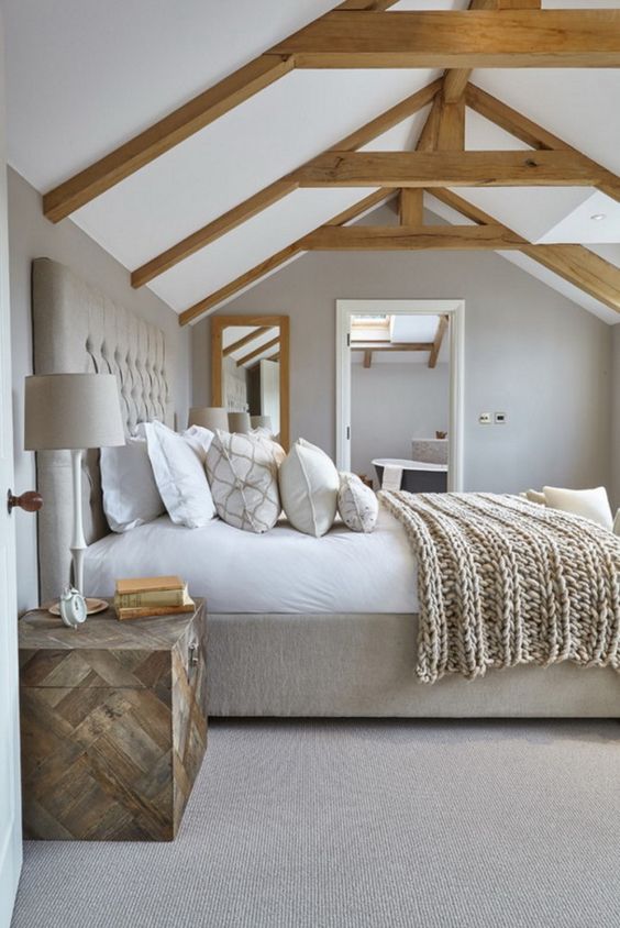 a neutral modern country bedroom done in grey, with wooden beams and an upholstered bed, a wooden chest and neutral bedding