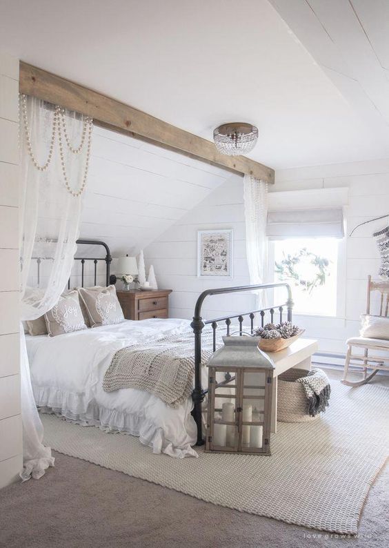 a neutral modern country bedroom with white planked walls, a wooden beam over the metal bed, neutral furniture and a large candle lantern