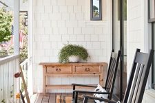 a pretty and simple farmhouse porch with a vintage console table, black rockers and printed pillows, potted greenery and some lanterns is welcoming