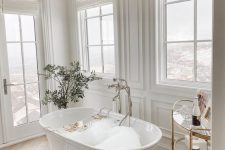 a refined and chic fancy bathroom with white paneled walls, windows, a luxurious bathtub, a crystal chandelier and a potted plant