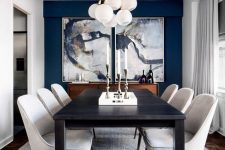 a refined and elegant dining room with a navy accent wlal, beautiful artworks, a long black table, creamy chairs and a cluster of pendant lamps