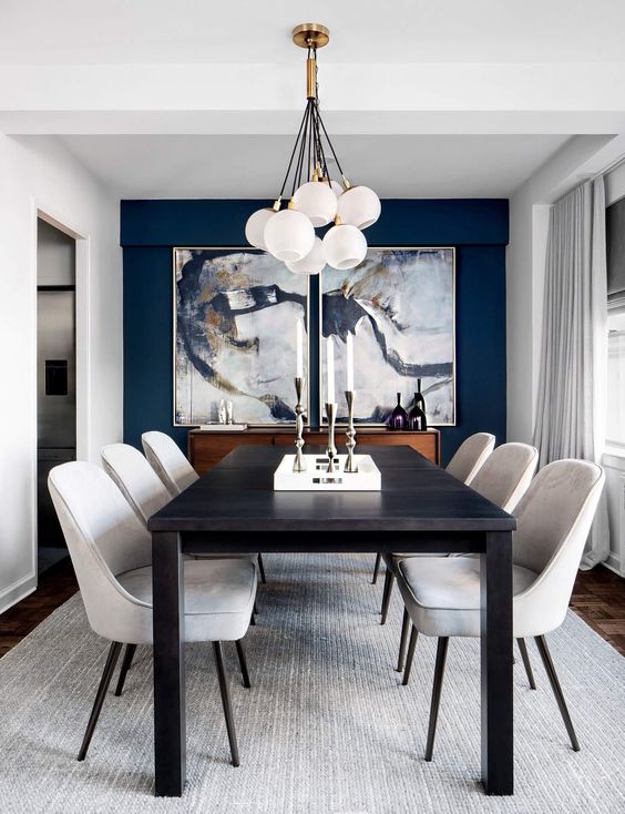 a refined and elegant dining room with a navy accent wlal, beautiful artworks, a long black table, creamy chairs and a cluster of pendant lamps