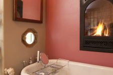 a refined bathroom with a pink accent wall and a built-in fireplace, a black vintage bathtub, a sculpture, a mirror in an ornated frame, a sconce and a mosaic tile floor