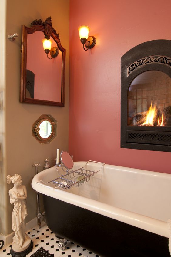 A refined bathroom with a pink accent wall and a built in fireplace, a black vintage bathtub, a sculpture, a mirror in an ornated frame, a sconce and a mosaic tile floor