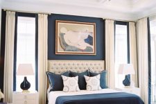 a refined bedroom with a navy accent wall, a creamy upholstered bed, navy and white bedding, creamy curtains and white nightstands