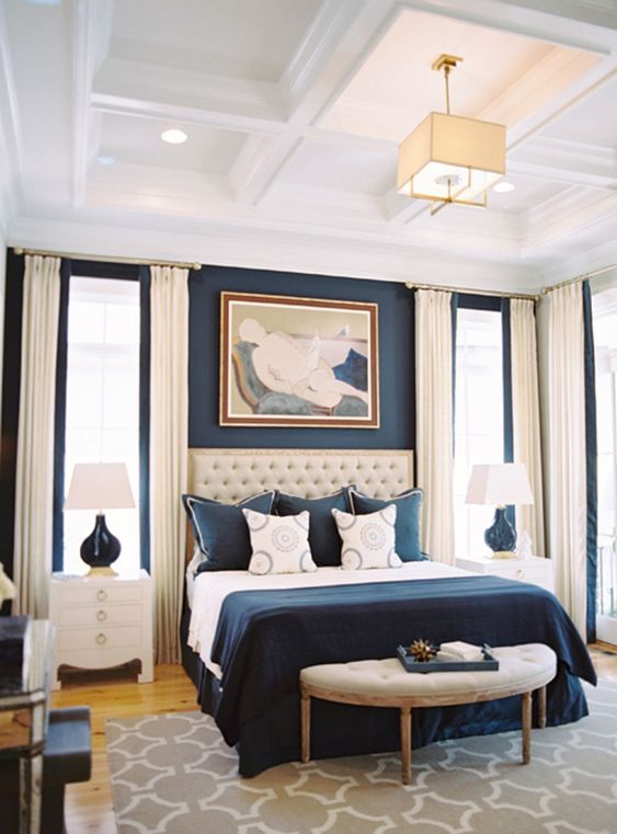 a refined bedroom with a navy accent wall, a creamy upholstered bed, navy and white bedding, creamy curtains and white nightstands