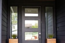 a simple and laconic modenr entrance with black walls and a glazed door, tall wooden planters with greenery and a printed rug