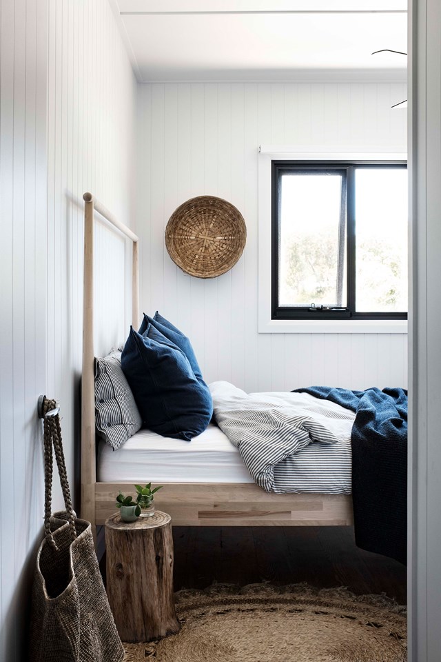 a small bedroom with white planked walls, a wooden bed, a jute rug, a tree stump side table, a decorative basket and navy and striped bedding