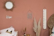 a small boho kid’s bedroom with a pink accent wall, simple wooden furniture and a peacock chair, a sunburst mirror and pampas grass