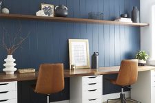 a stylish home office with a navy planked wall, a floating shelf and a double desk with a stained desktop, leather chairs and some pretty vases