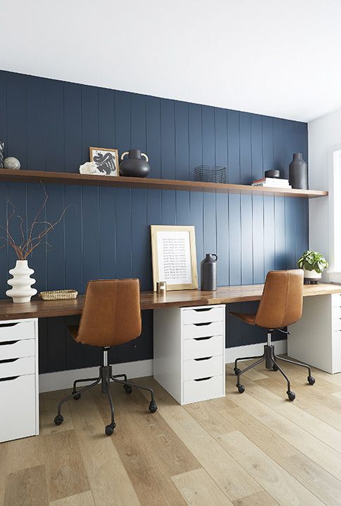 a stylish home office with a navy planked wall, a floating shelf and a double desk with a stained desktop, leather chairs and some pretty vases