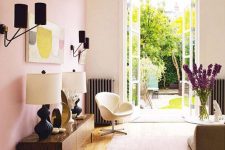 a stylish living room with a pink accent wall, some chic furniture in modern style, a catchy chandelier and black sconces