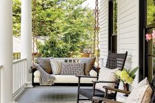 a stylish modern farmhouse porch with a black suspended daybed with pillows, black wooden rockers, a striped rug and some blooms