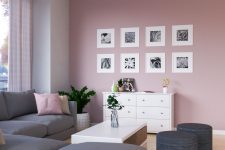 a stylish modern living room with a pale pink accent wall and a grid gallery wall, a white sideboard, a grey sectional, a low white coffee table
