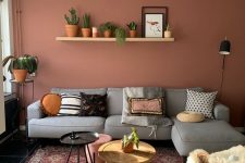 a stylish modern living room with a pink accent wall, a grey sectional, a printed boho rug, a shelf with lots of potted plants and an arrangement of side tables