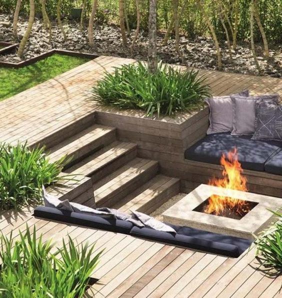 A sunken patio with a deck, a fire pit, built in benches with pillows and grasses growing around is a very lovely space
