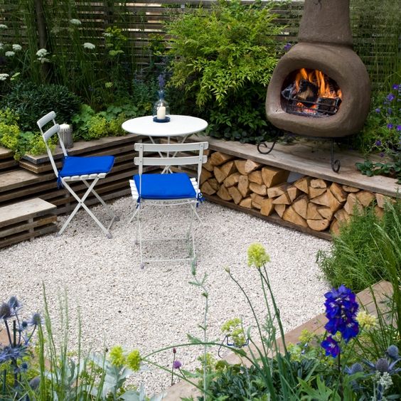 a sunken patio with gravel on the ground, a hearth, a small dining space and lots of greenery and blooms growing around