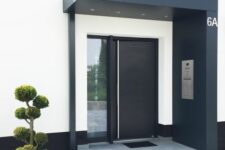 a super chic modern entrance with a concrete ladder, a black over with lights, a black door and greenery next to the ladder