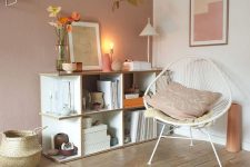 a welcoming Scandinavian nook with an open storage unit, a white chair, some lamps, a pale pink accent wall and a basket