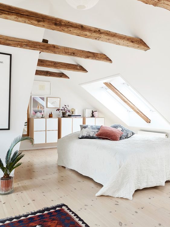 a welcoming attic bedroom with wooden beams, floating dressers and a bed by the skylight, a bright rug and pillows and a potted plant
