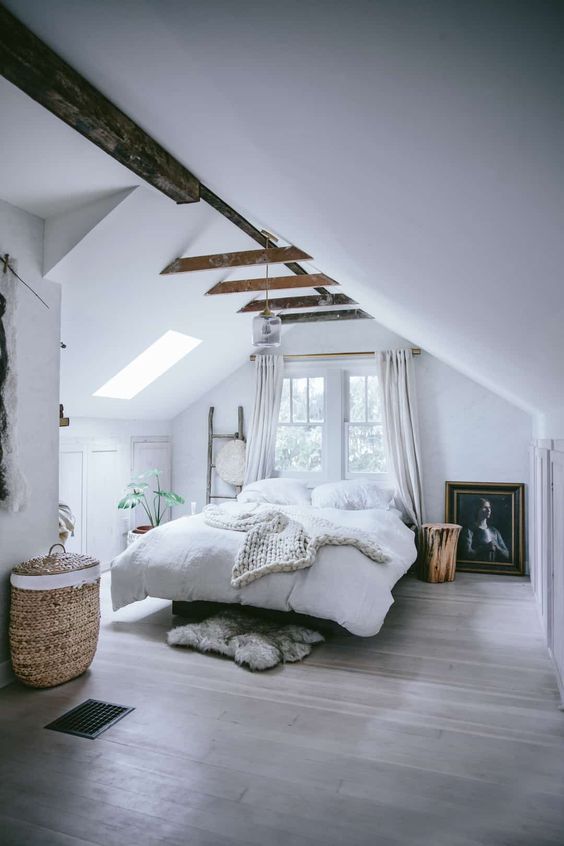 a welcoming modern country bedroom with an attic ceiling and wooden beams, a bed, a window and a skylight, potted plants and a basket