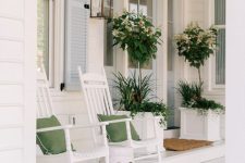 a white farmhouse porch with rockers and green pillows, a side table, a lantern and lots of greenery in large white planters is chic and clean