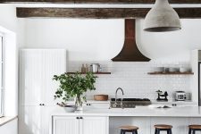 a white modern country kitchen with white planked cabinets, dark ceiling beams, pendant lamps, tall stools and shelves