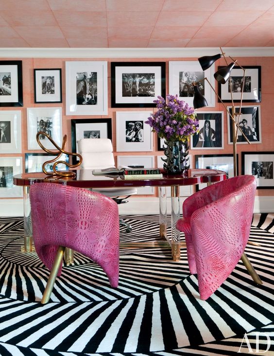 an artistic home office with a pink accent wall fully covered with a black and white gallery wall, a unique desk and hot pink leather chairs on brass legs