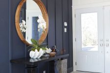an elegant coastal farmhouse entryway with a navy paneled wall, a tiled floor, a black console table, a round mirror in a wooden frame and a vintage chandelier