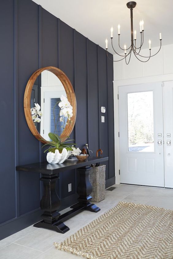 an elegant coastal farmhouse entryway with a navy paneled wall, a tiled floor, a black console table, a round mirror in a wooden frame and a vintage chandelier