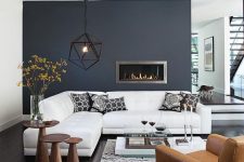 an elegant living room with a navy accent wall, a white corner sofa, a tan leather chair and an arrangement of wooden side tables plus a built-in fireplace