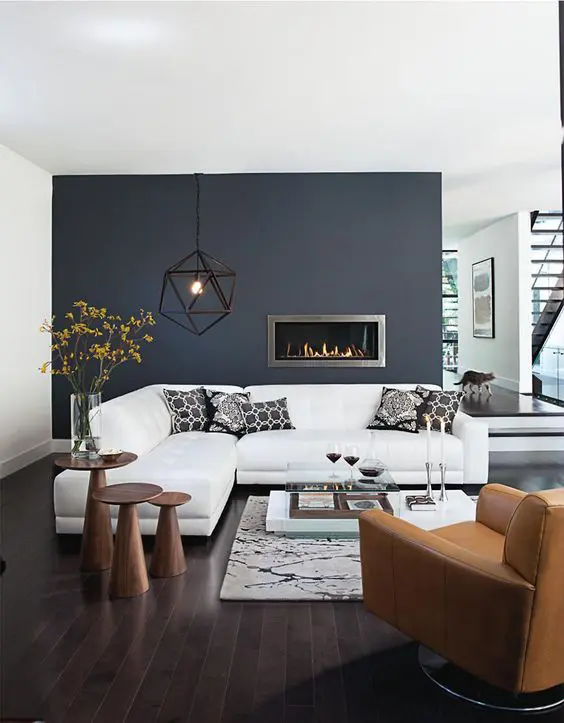 An elegant living room with a navy accent wall, a white corner sofa, a tan leather chair and an arrangement of wooden side tables plus a built in fireplace