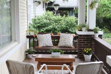 an elegant modern farmhouse porch with a dark stained suspended bench and pillows, white chairs, a wooden coffee table and potted greenery and blooms