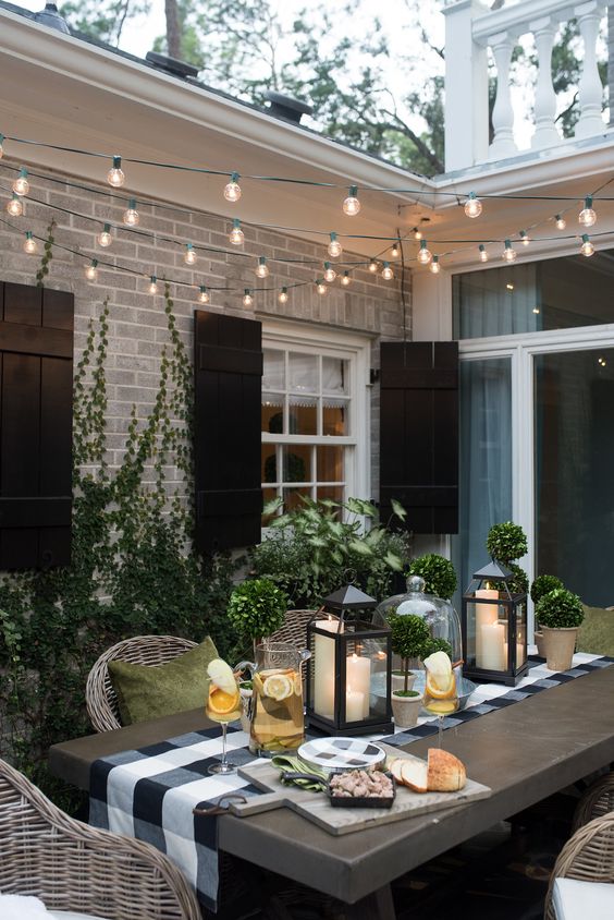 an inviting modern country patio with a table and wicker chairs, muted color pillows, potted greenery and candle lanterns plus lights over the space