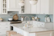 a lovely modern coastal kitchen with white cabinets, a blue subway tile backsplash, woven stools and elegant bubble pendant lamps