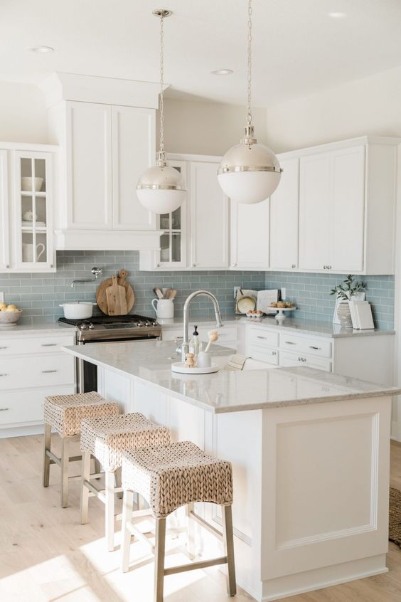 a lovely modern coastal kitchen with white cabinets, a blue subway tile backsplash, woven stools and elegant bubble pendant lamps