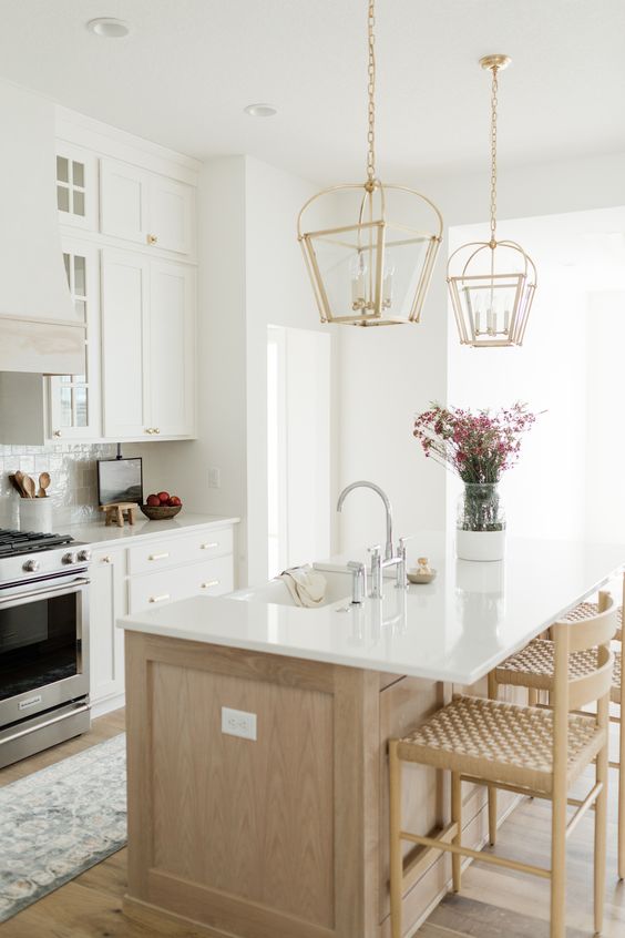 an elegant modern farmhouse kitchen with white cabinets, a light stained kitchen island, gold pendant lamps and woven chairs