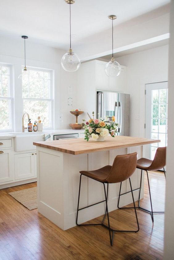 a pretty white farmhouse kitchen with shaker style cabinets, a small kitchen island with a butcherblock countertop and leather stools