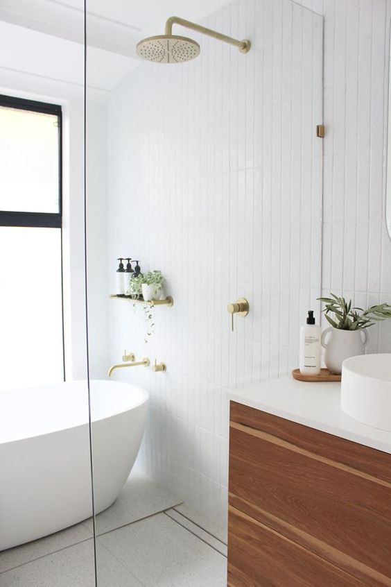a beautiful minimalist bathroom with white skinny tiles, a chic tub, gold appliances, a wooden vanity and potted greenery is chic